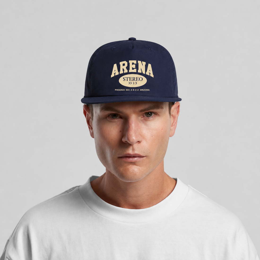 A man wearing a navy blue snapback cap with cream-colored 'ARENA STEREO' text on the front, paired with a white crewneck t-shirt against a light gray background.