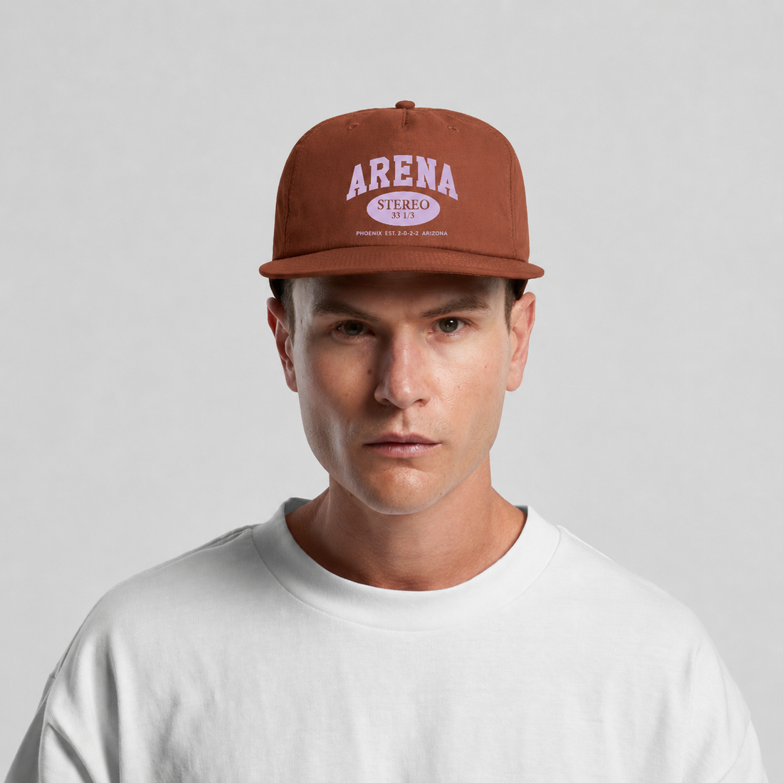 A man wearing a rust-brown flat-brim cap with 'ARENA STEREO' printed in white on the front, paired with a white crewneck t-shirt, against a light gray studio background.
