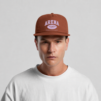 A man wearing a rust-brown flat-brim cap with 'ARENA STEREO' printed in white on the front, paired with a white crewneck t-shirt, against a light gray studio background.