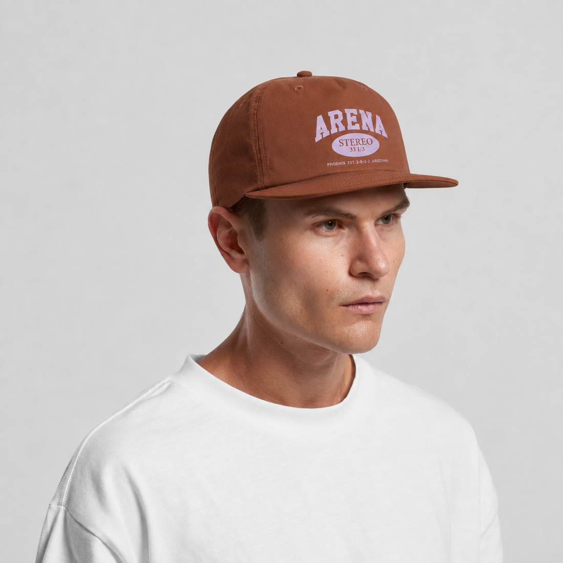 A man wearing a brown snapback cap with a white 'ARENA' logo, paired with a white t-shirt, photographed against a light gray studio background.