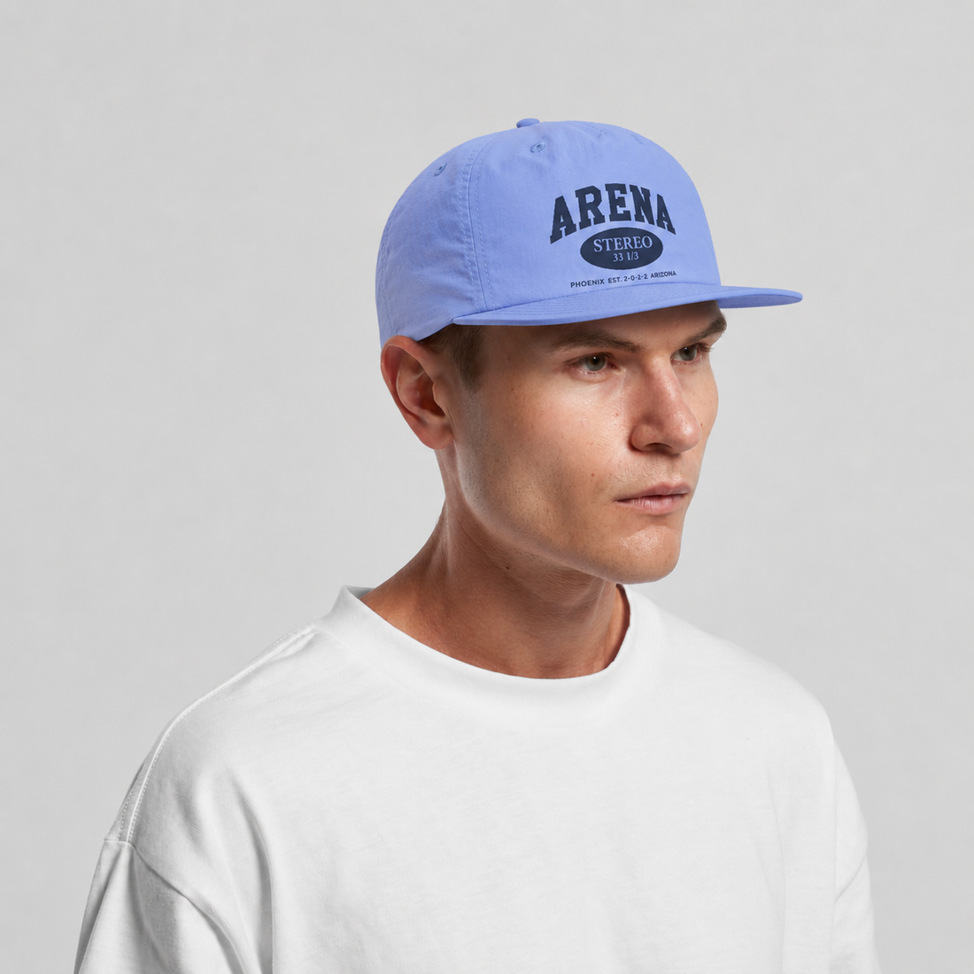 A man wearing a light blue snapback cap with 'ARENA STEREO' printed in dark blue on the front, paired with a white crewneck sweatshirt, set against a plain light gray background.