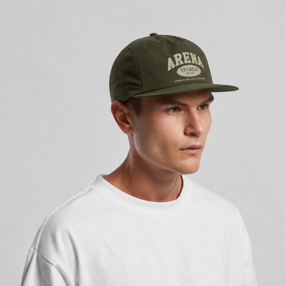 A man wearing an olive green flat-brim cap with 'ARENA STEREO' printed in cream on the front, paired with a white crewneck t-shirt, set against a plain light gray background.