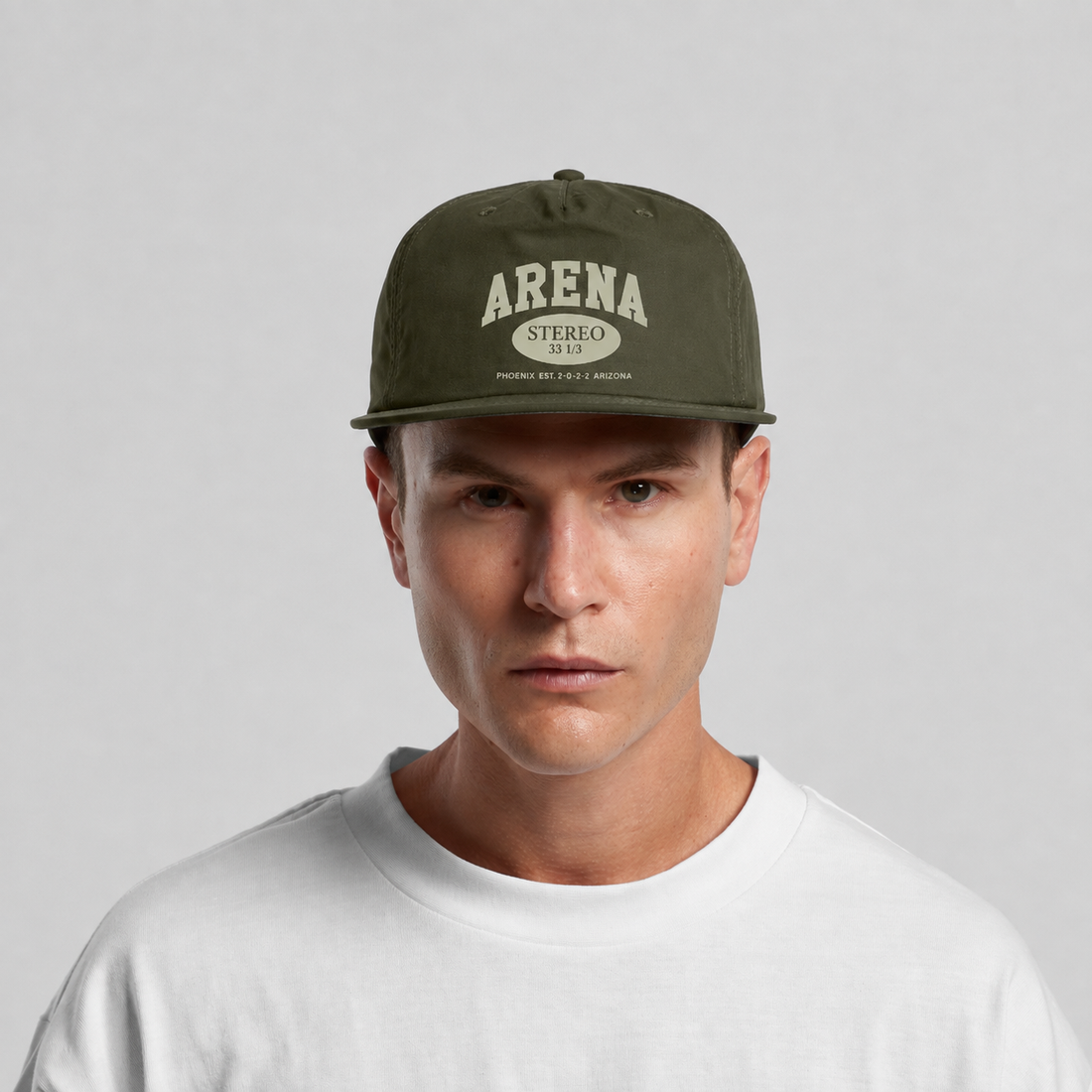 A man wearing an olive green flat-brim cap with 'ARENA STEREO' printed in cream on the front, paired with a white crewneck t-shirt, set against a plain light gray background.