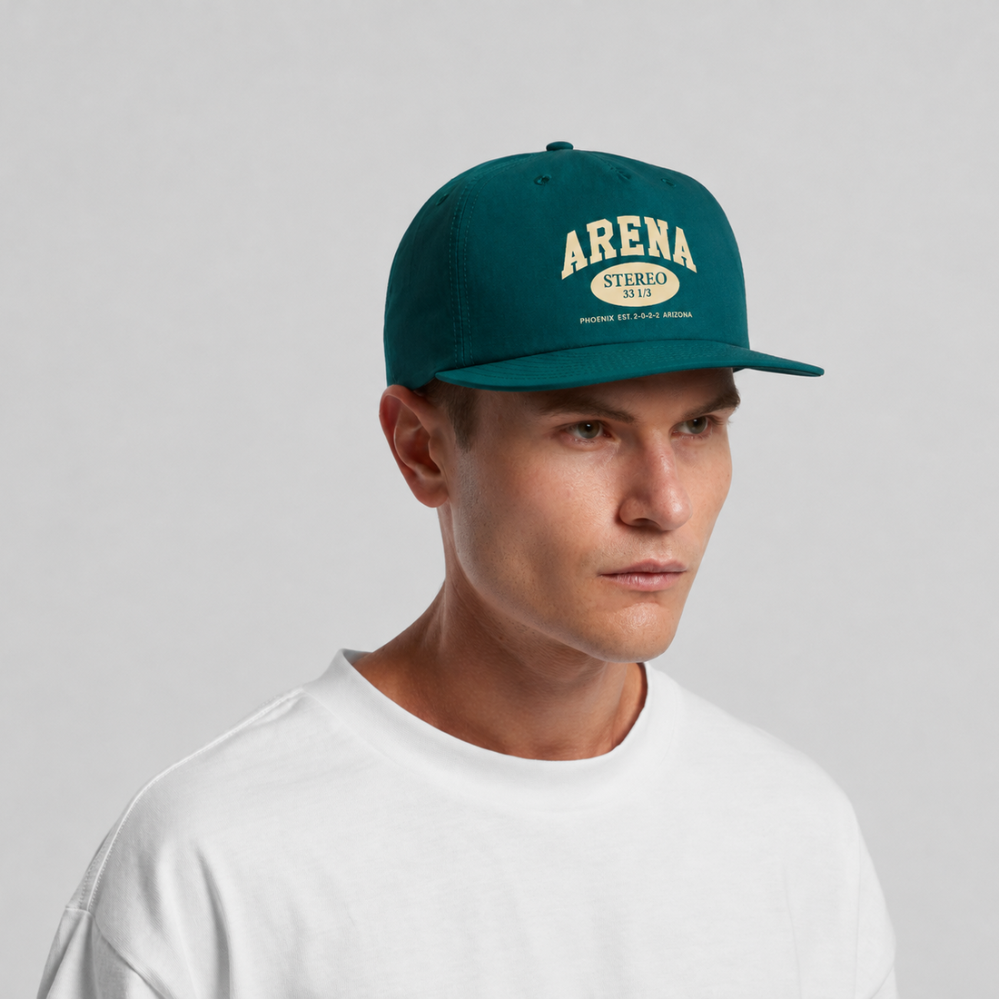 A man wearing a dark teal green snapback cap with 'ARENA STEREO' printed in cream on the front, paired with a white t-shirt against a light gray studio background.