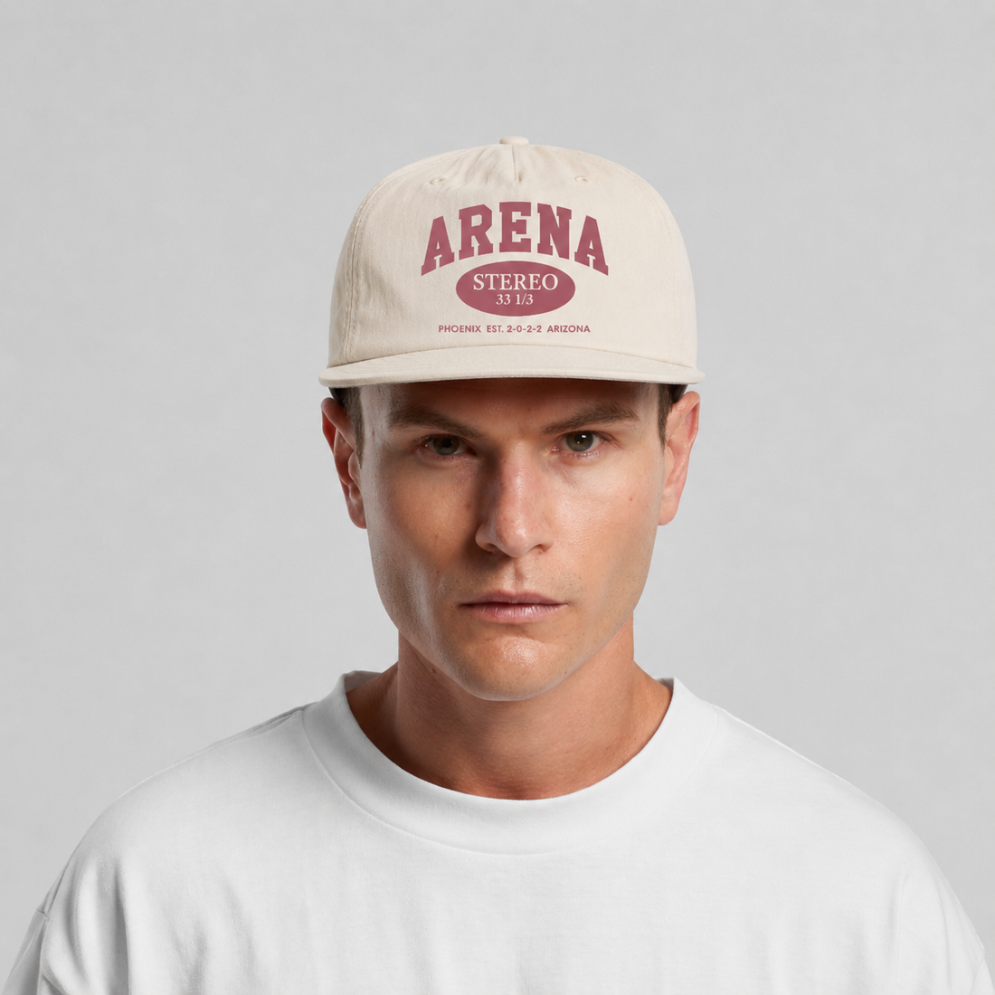 A man wearing a bone-colored surf cap with a rose 'ARENA' graphic on the front, paired with a white t-shirt against a light gray background.