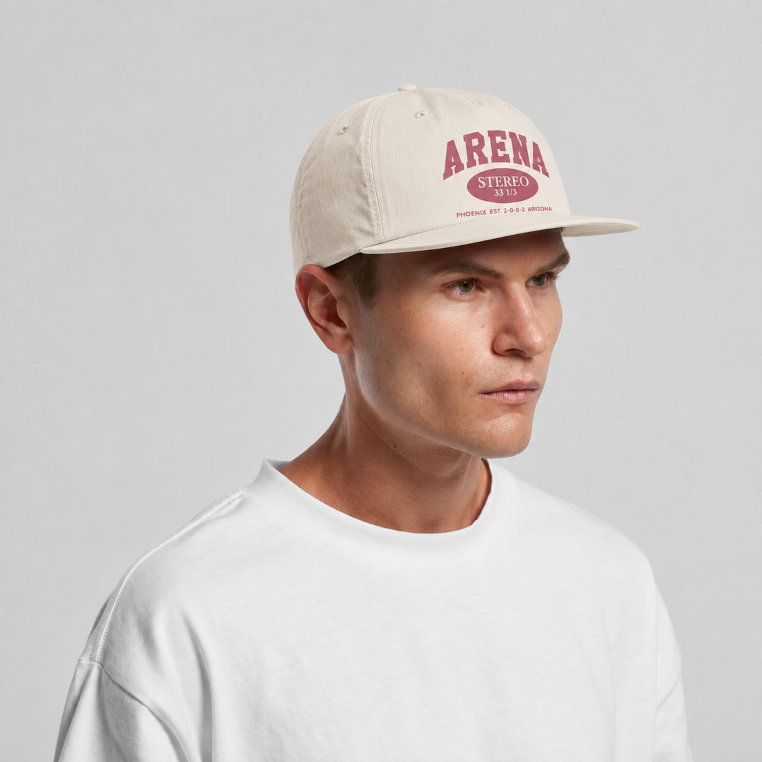 A man wearing a cream-colored surf cap with a rose 'ARENA' logo on the front, paired with a white crewneck t-shirt, against a light gray studio background.