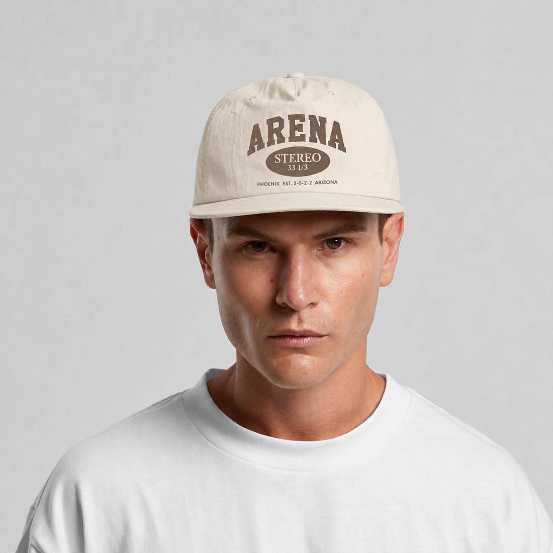A man wearing a bone colored surf cap with 'ARENA STEREO' printed in brown on the front, paired with a white t-shirt against a light gray background.