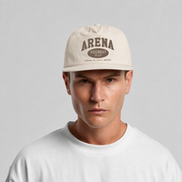 A man wearing a bone colored surf cap with 'ARENA STEREO' printed in brown on the front, paired with a white t-shirt against a light gray background.