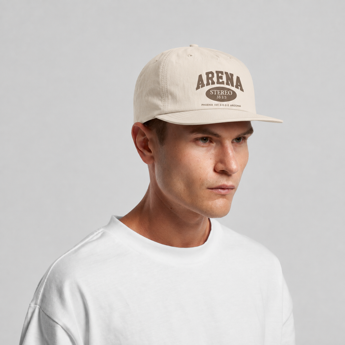 A man wearing a bone colored snapback cap with a brown 'ARENA' logo, paired with a white t-shirt, against a light gray studio background.