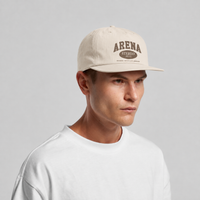 A man wearing a bone colored snapback cap with a brown 'ARENA' logo, paired with a white t-shirt, against a light gray studio background.
