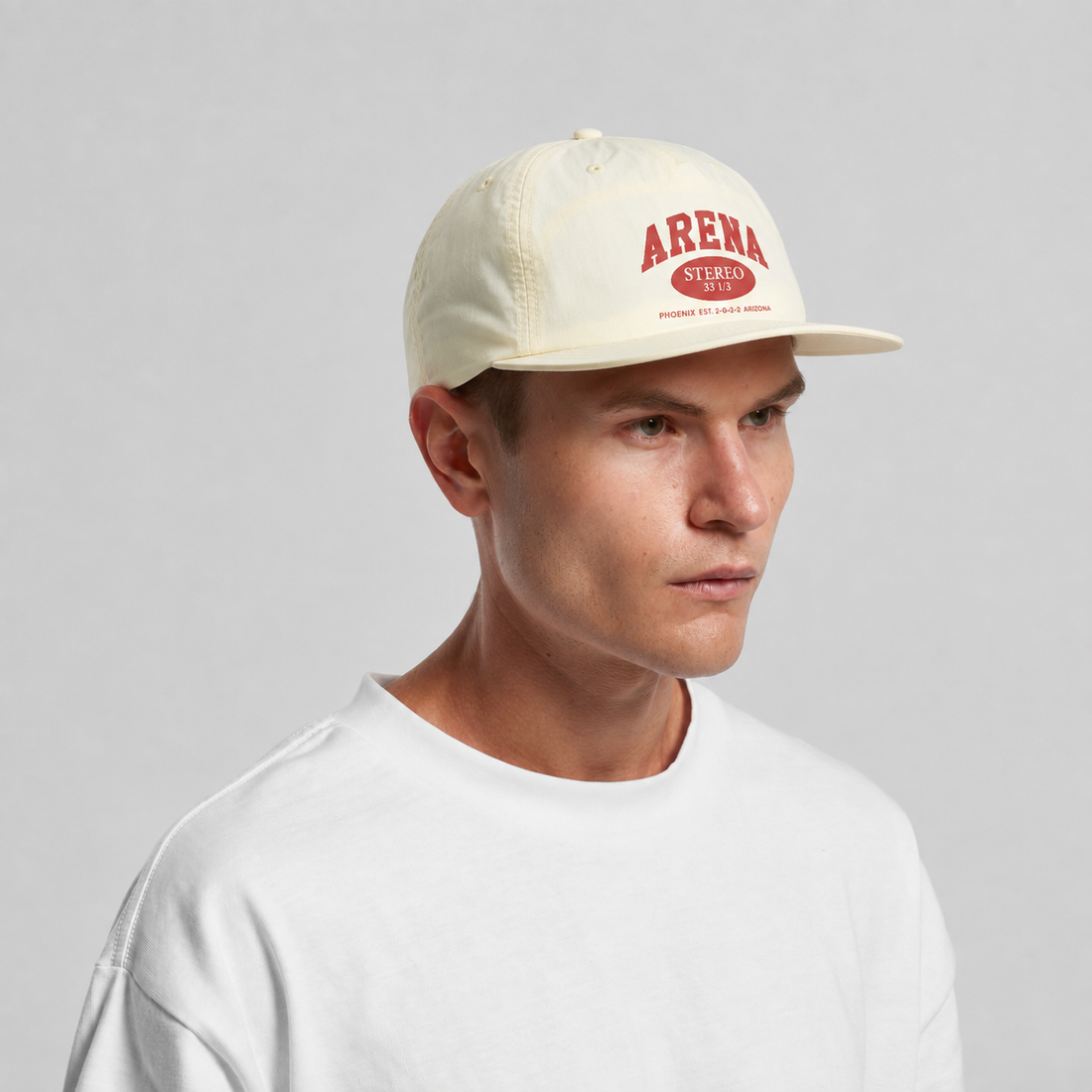 A man wearing a cream-colored snapback cap with a red 'ARENA' logo on the front, paired with a white t-shirt, against a plain light gray background.