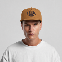 A man wearing a tan snapback cap with a dark brown 'ARENA STEREO' logo on the front, paired with a white crewneck t-shirt, against a light gray background.