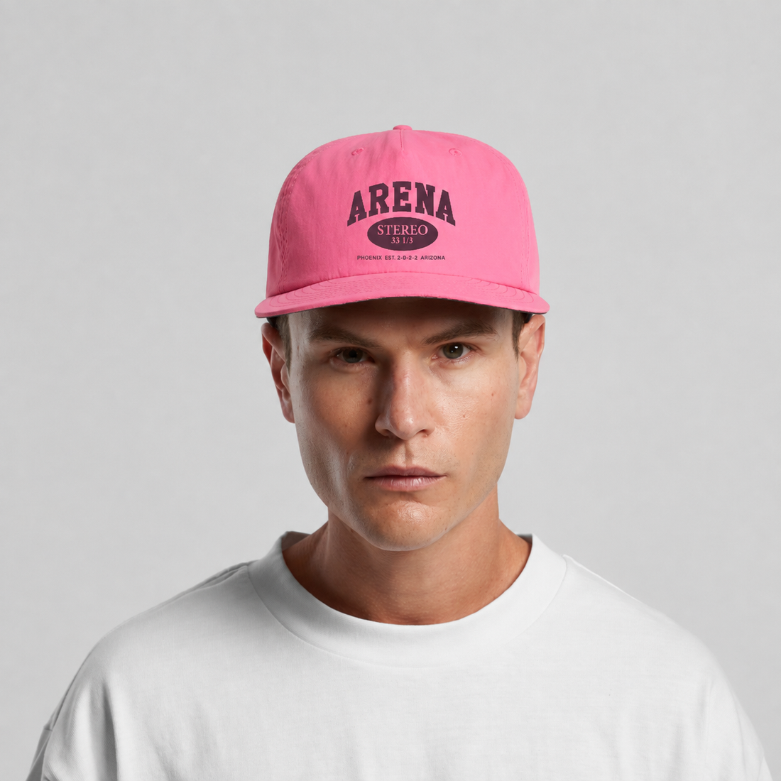 A man wearing a bright pink baseball cap with 'ARENA STEREO' printed in black on the front, paired with a white t-shirt against a light gray background.