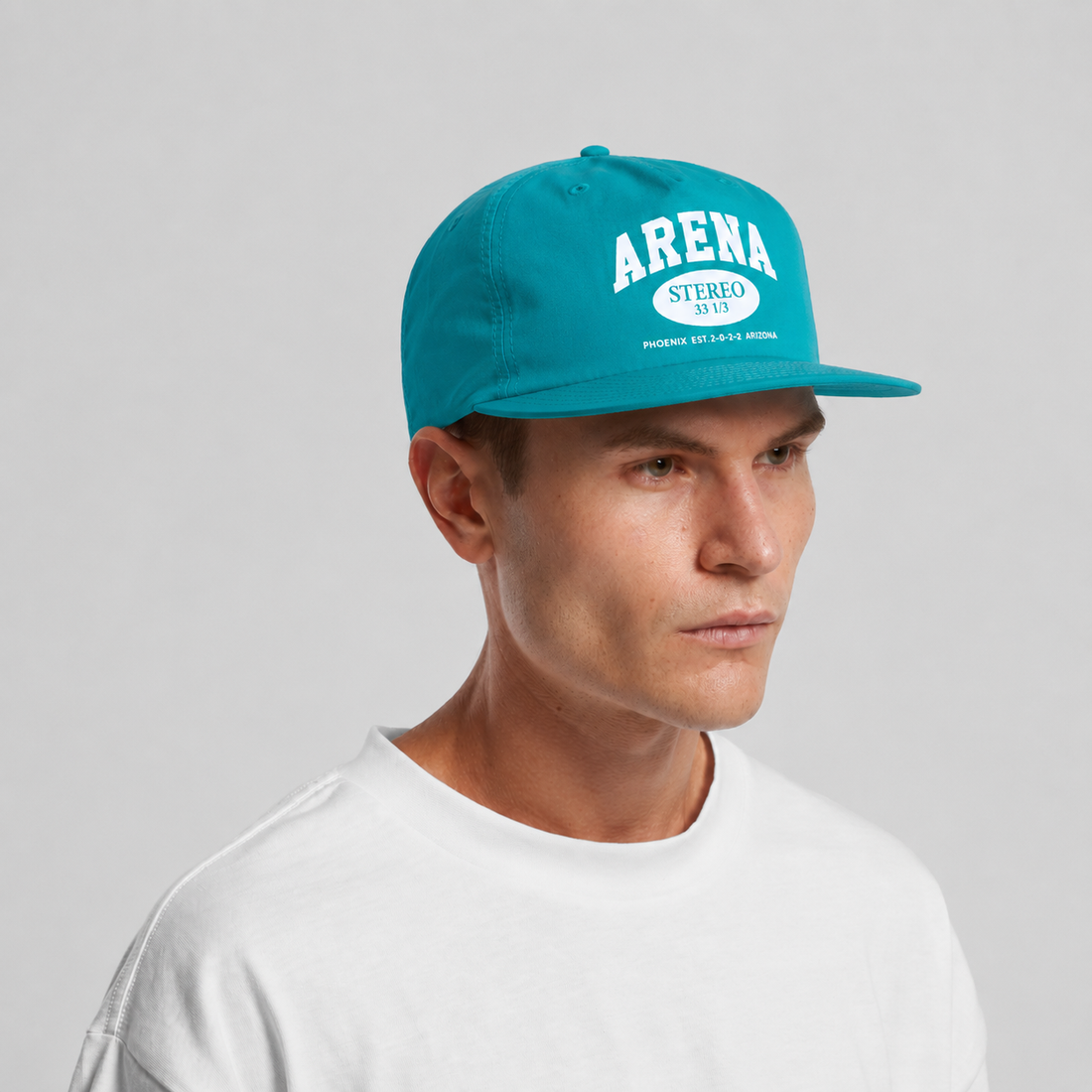 A man wearing a teal blue flat-brim surf cap with white 'ARENA STEREO' text on the front, paired with a white t-shirt, against a light gray studio background.