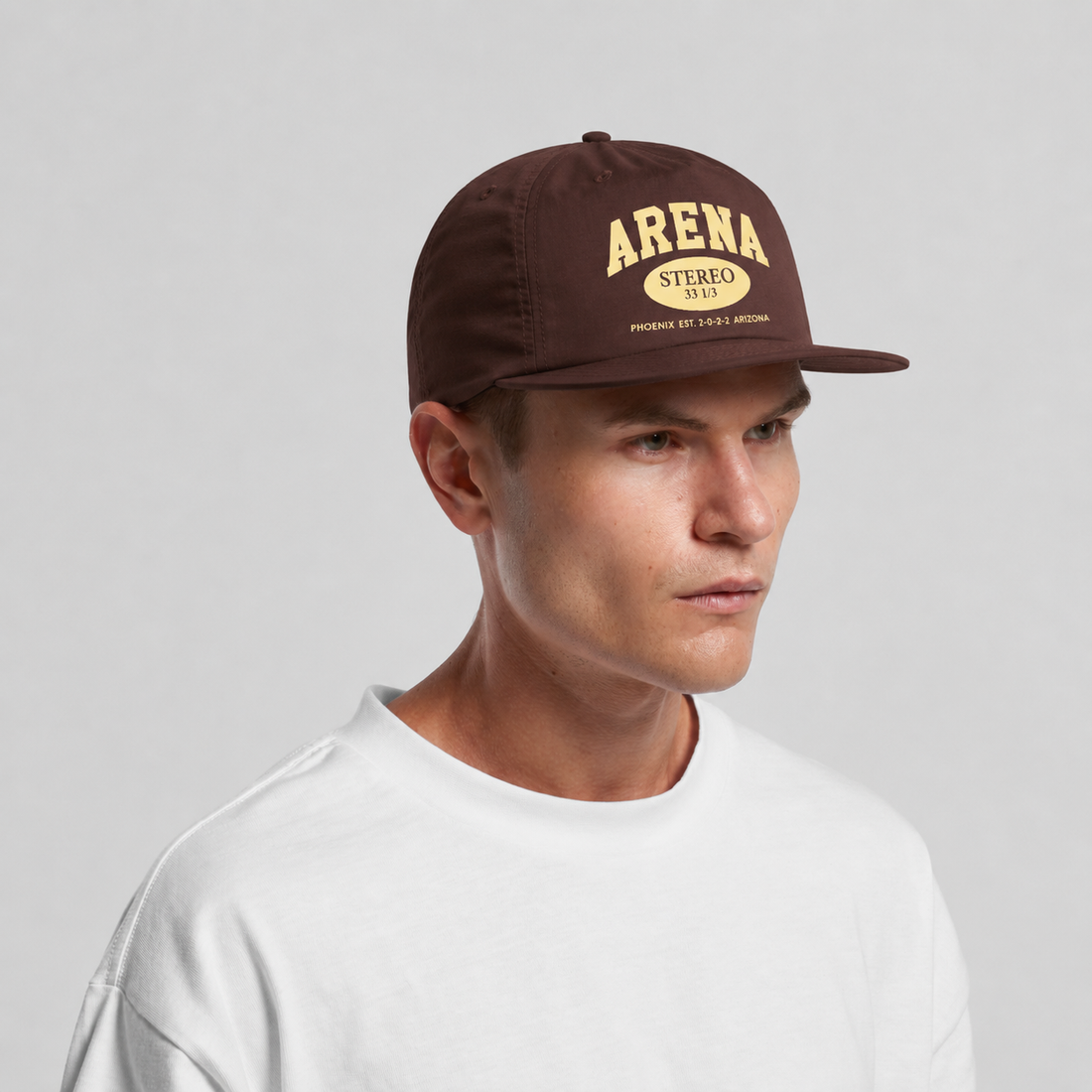 A man wearing a chestnut brown snapback cap with a gold 'ARENA STEREO' logo on the front, paired with a white t-shirt, against a light gray studio background.