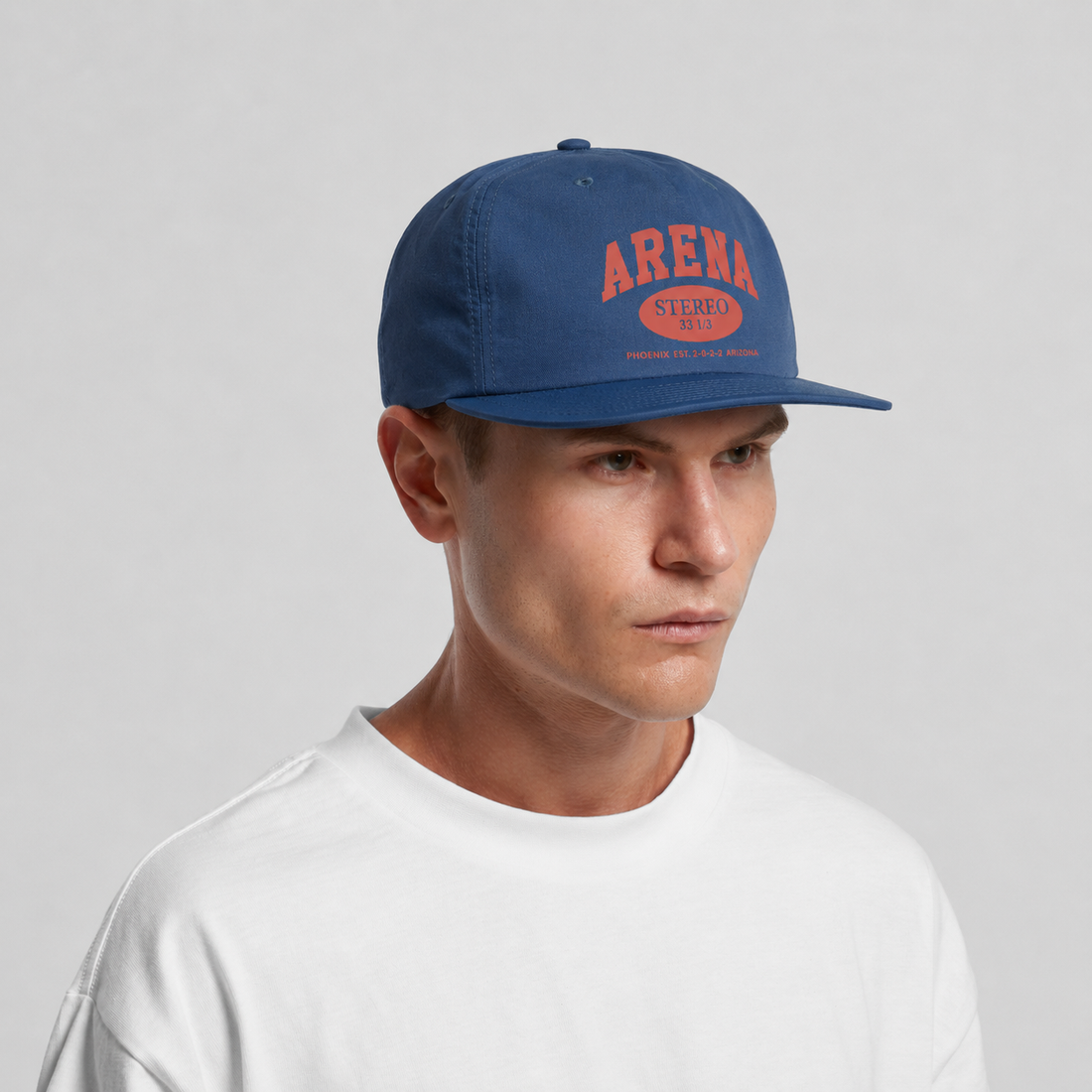 A man wearing a blue snapback cap with a red 'ARENA STEREO' logo on the front, paired with a white t-shirt, photographed against a neutral light gray background.