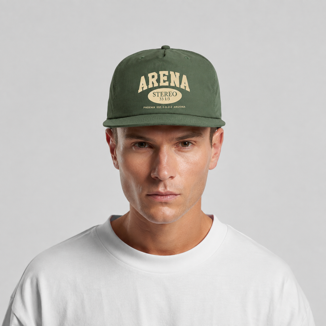 A man wearing a cypress green flat-brim cap with a cream-colored 'ARENA STEREO' logo on the front, paired with a white crewneck t-shirt, against a light gray studio background.