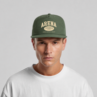 A man wearing a cypress green flat-brim cap with a cream-colored 'ARENA STEREO' logo on the front, paired with a white crewneck t-shirt, against a light gray studio background.