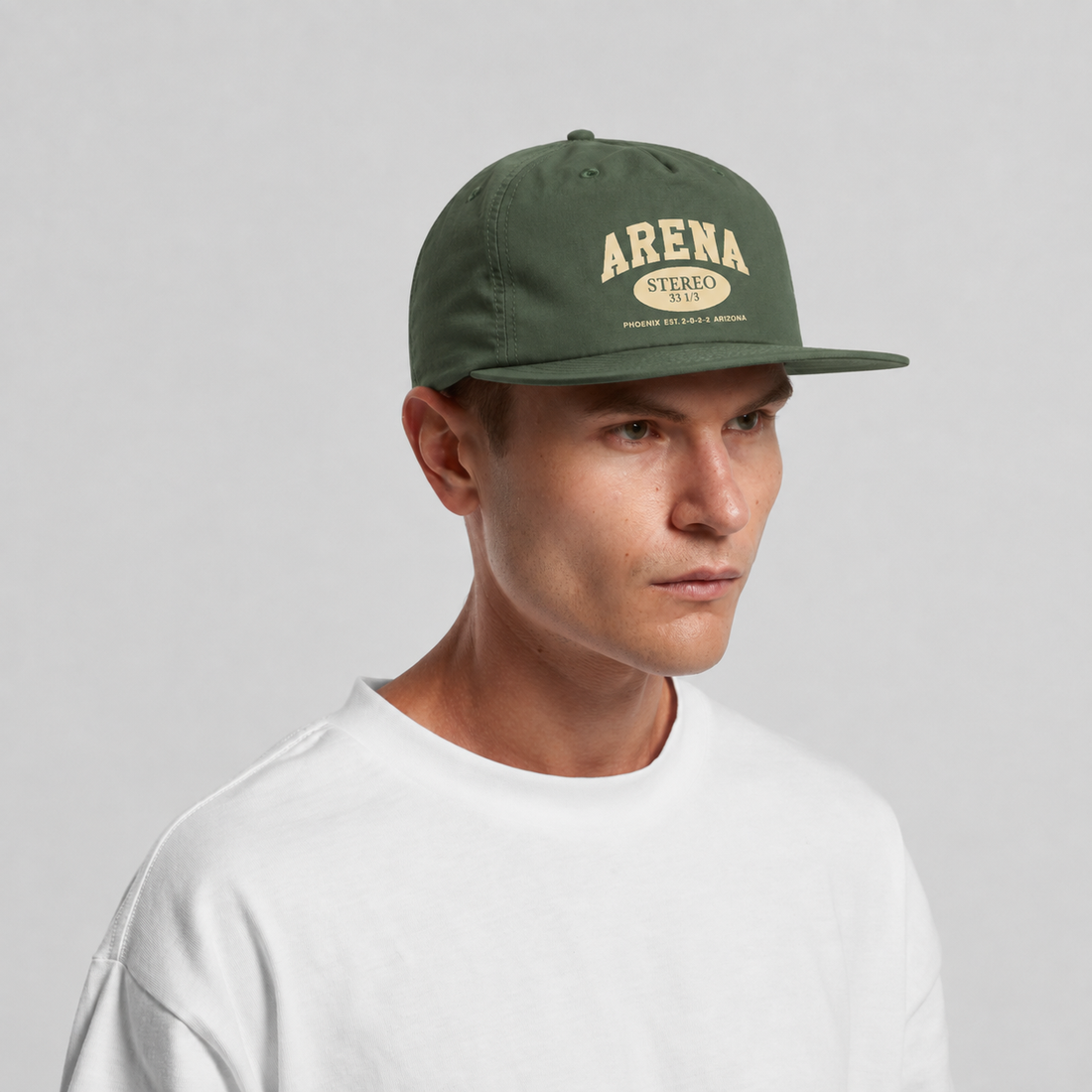 A man wearing a cypress green flat-brim cap with 'ARENA STEREO' printed in cream on the front, paired with a white crewneck t-shirt, set against a plain light gray background.