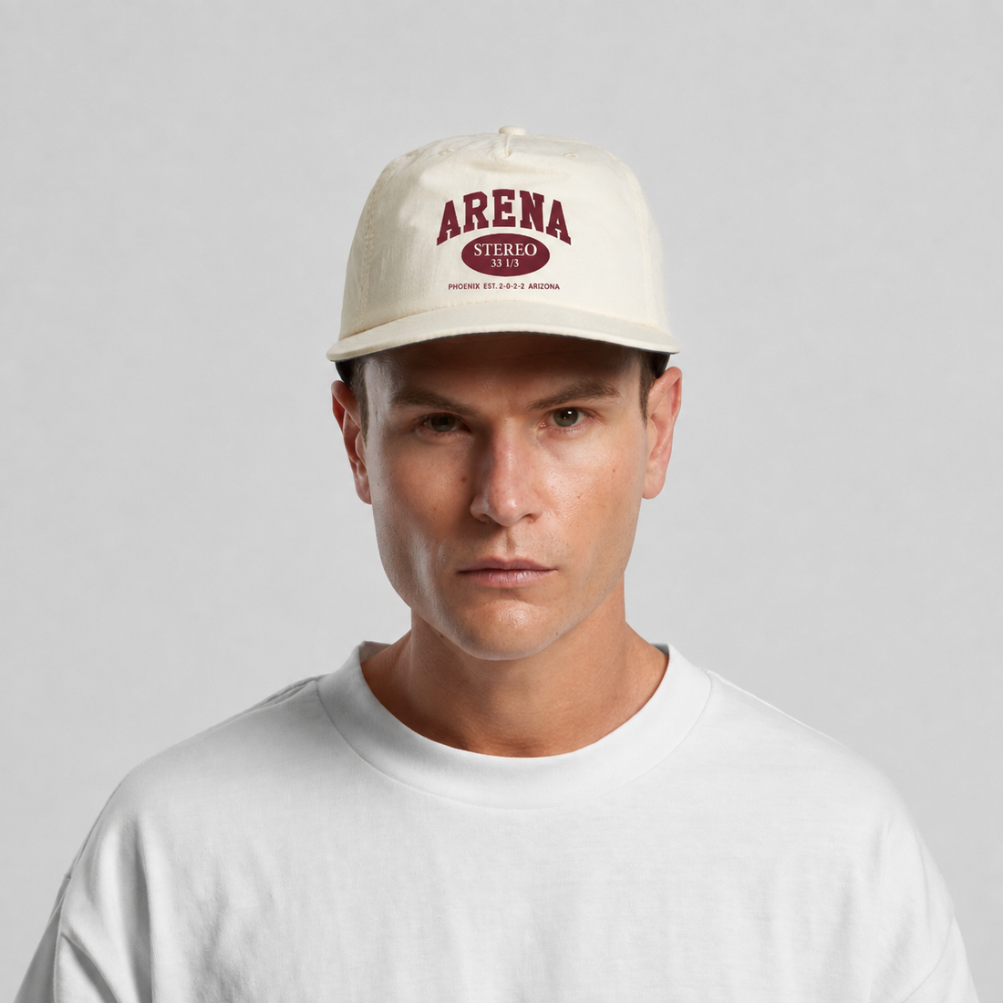A man wearing an ecru-colored surf cap with a maroon 'ARENA STEREO' logo on the front, paired with a white crewneck t-shirt, against a light gray studio background.