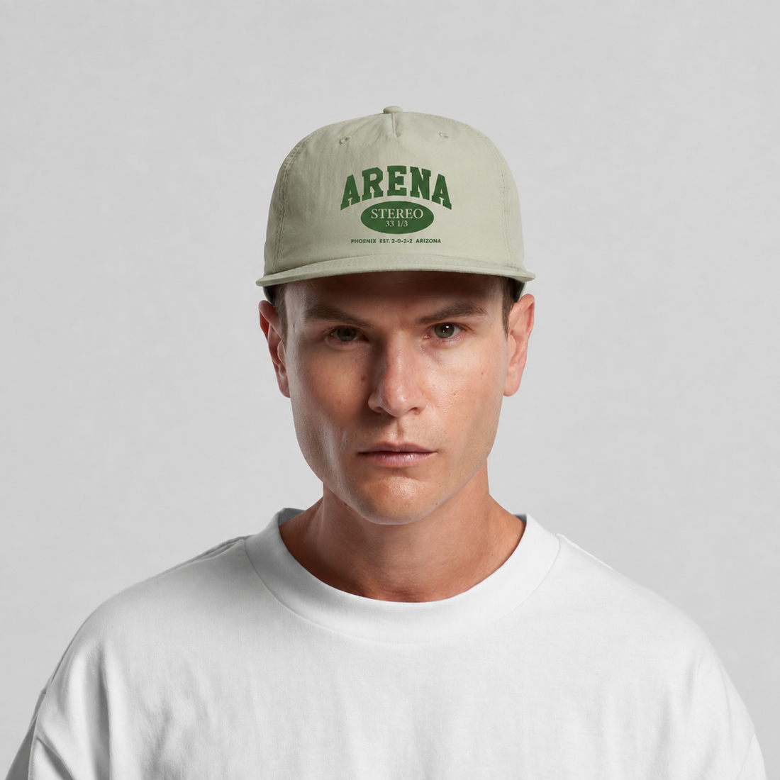 A man wearing a light sage green baseball cap with green 'ARENA' branding, paired with a white crewneck t-shirt, against a neutral light gray background.