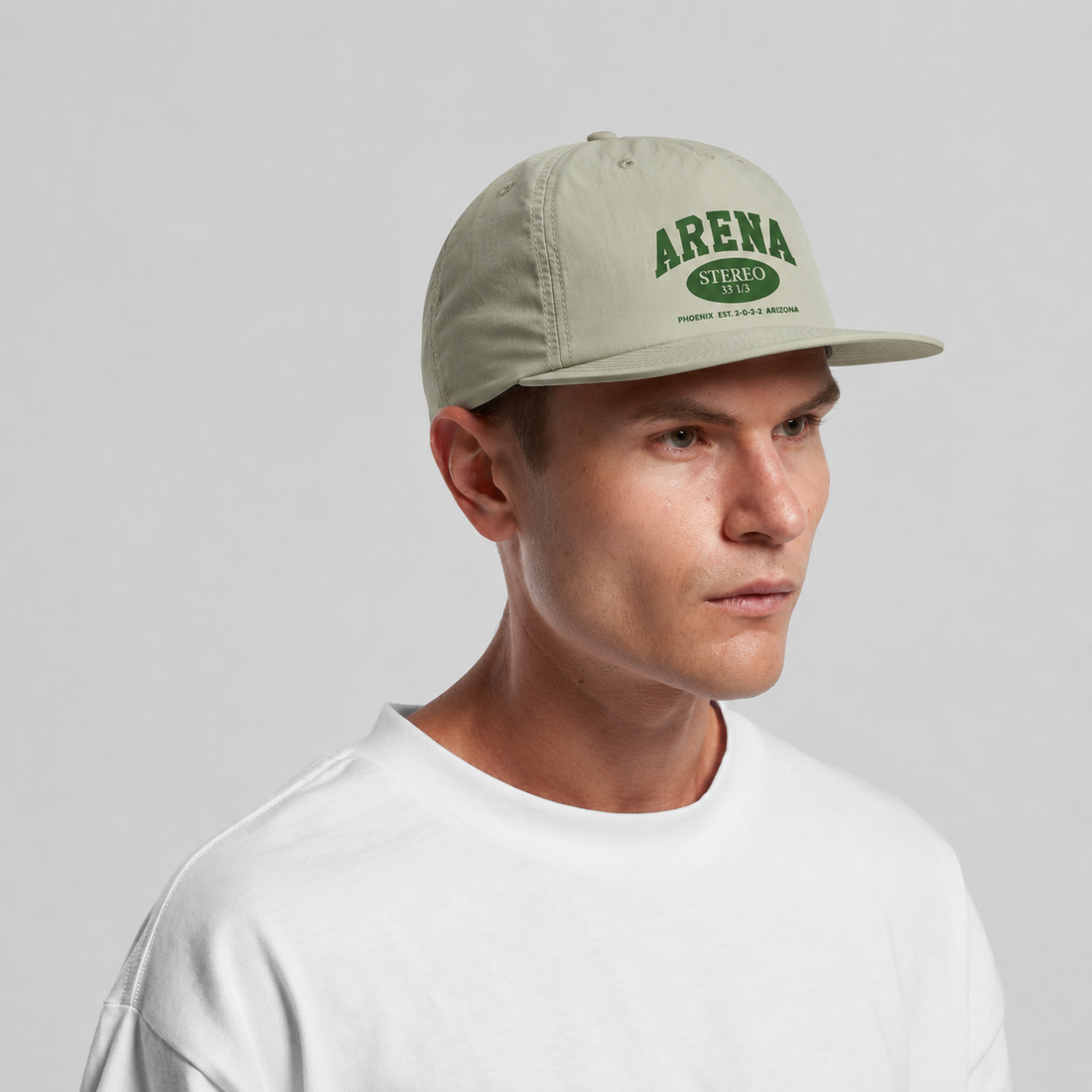 A man wearing a light sage green baseball cap with green 'ARENA' branding, paired with a white t-shirt, against a neutral gray background.