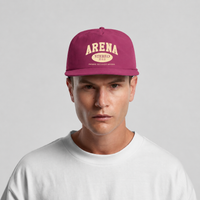 A man wearing a magenta snapback cap with a cream-colored 'ARENA STEREO' logo on the front, paired with a white t-shirt against a light gray background.