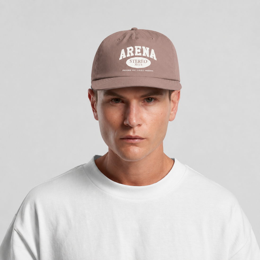 A man wearing a taupe brown baseball cap with 'ARENA STEREO' printed in white on the front, paired with a white crewneck t-shirt against a light gray background.