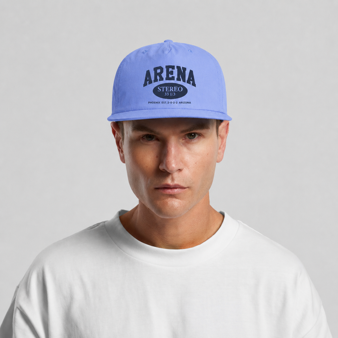 A man wearing a light blue snapback cap with 'ARENA STEREO' printed on the front, paired with a white t-shirt against a light gray background.