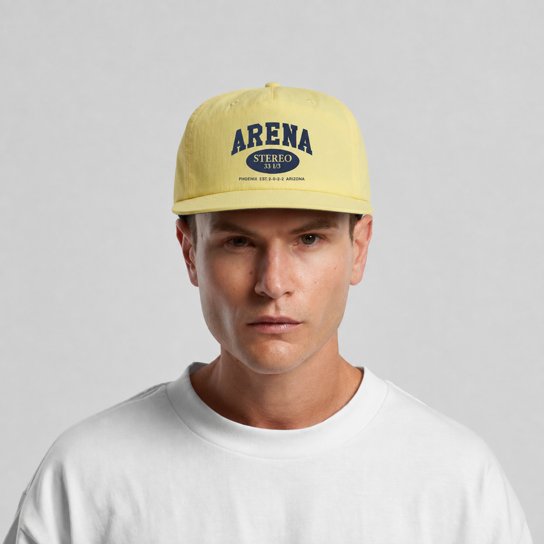 A man wearing a pale yellow baseball cap with a navy blue 'ARENA STEREO 1968' logo on the front, paired with a white t-shirt against a light gray background.