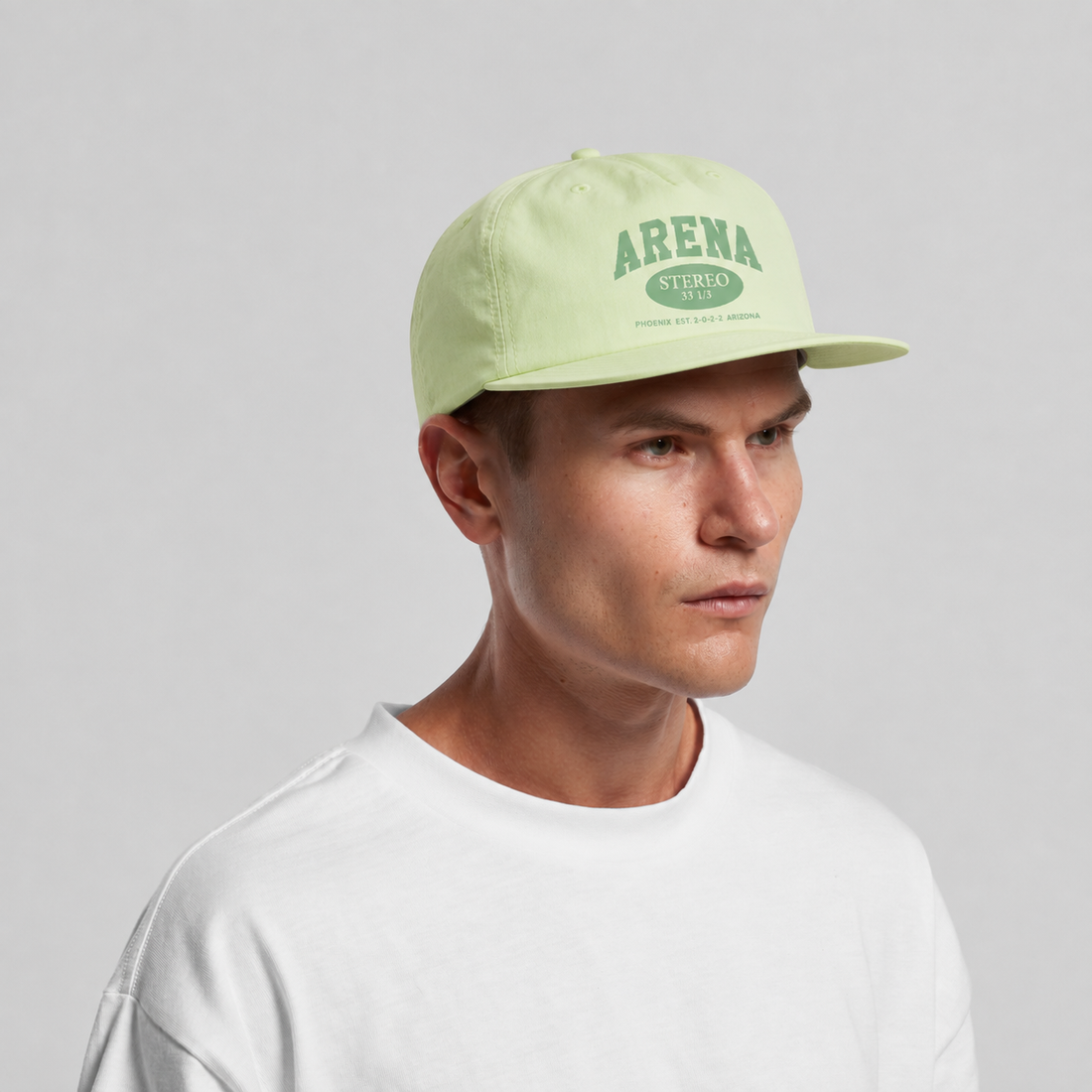 A man wearing a light yellow-green flat-brim cap with a green 'ARENA STEREO' logo, paired with a white t-shirt, photographed against a neutral light gray background.