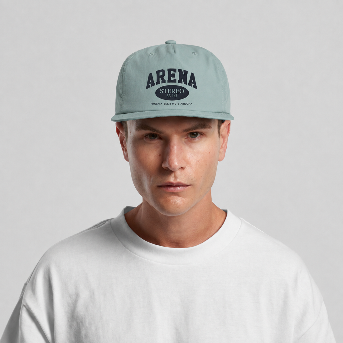 A man wearing a light blue snapback cap with 'ARENA STEREO' printed on the front, paired with a white t-shirt against a light gray background.