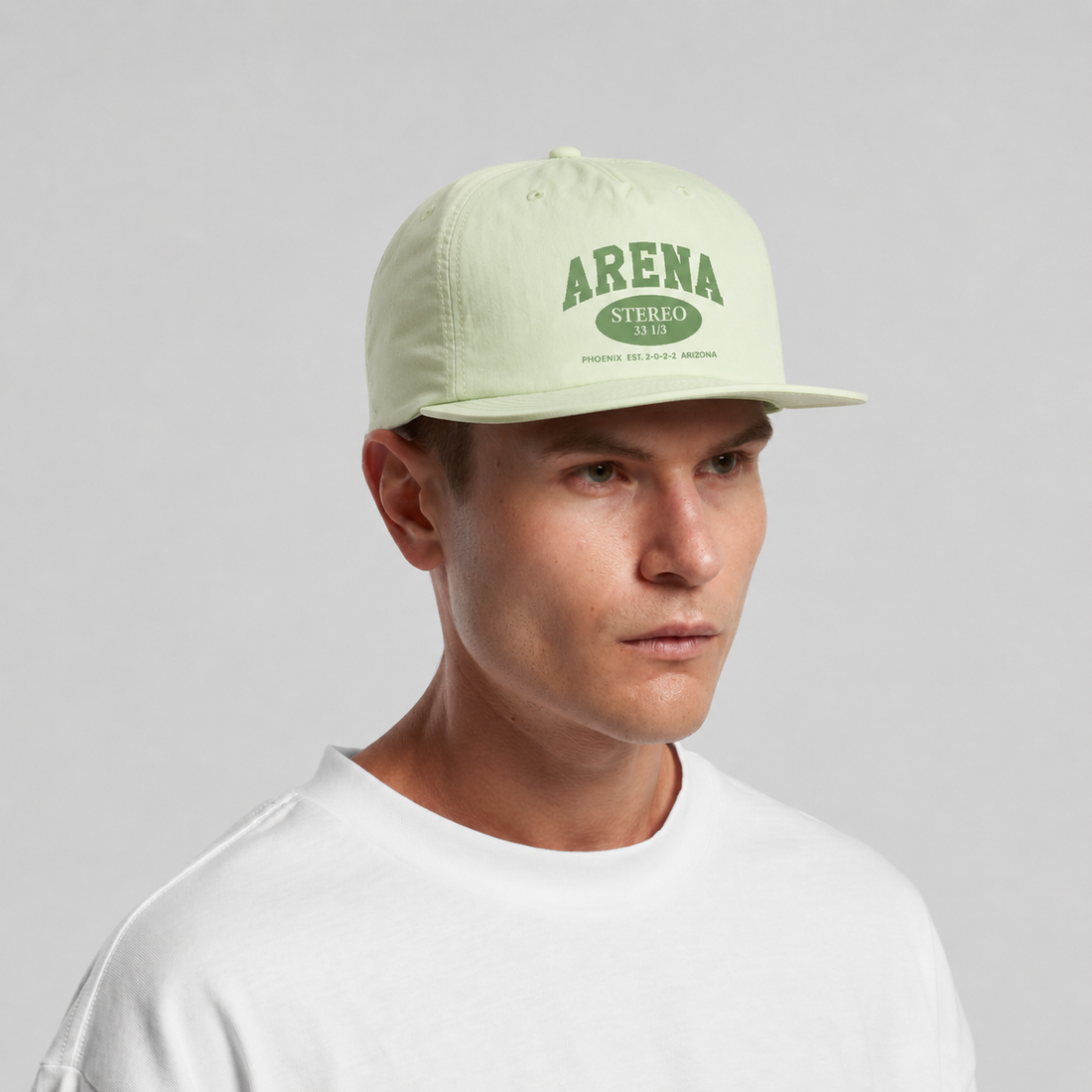 A man wearing a light sage green snapback cap with a dark green 'ARENA STEREO' logo on the front, paired with a white t-shirt against a neutral gray background.