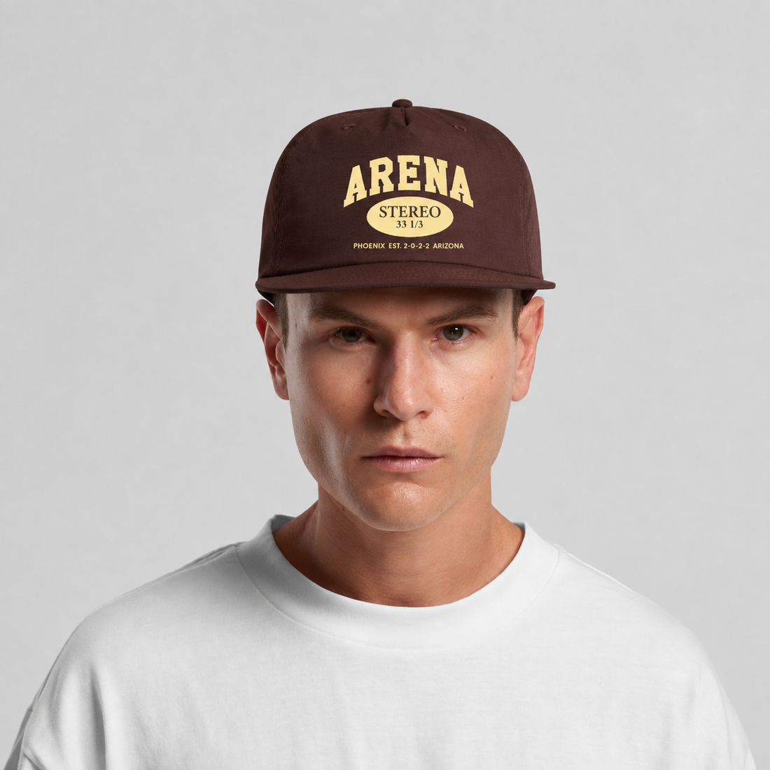 A man wearing a dark brown snapback cap with 'ARENA STEREO' printed in beige on the front, paired with a white t-shirt against a light gray background.