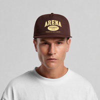 A man wearing a dark brown snapback cap with 'ARENA STEREO' printed in beige on the front, paired with a white t-shirt against a light gray background.