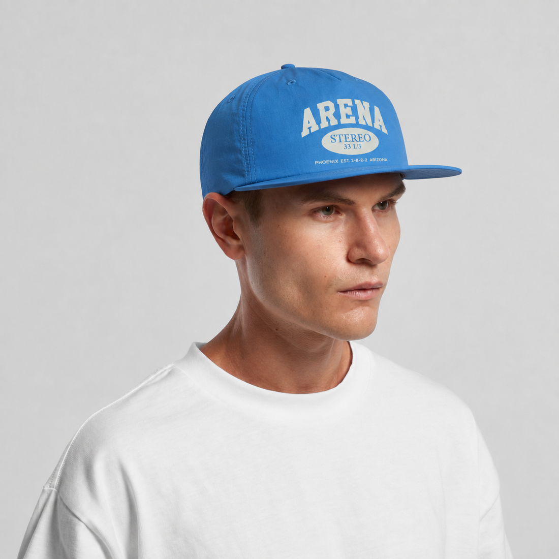 A man wearing a bright blue snapback cap with 'ARENA STEREO' printed in white on the front, paired with a white crewneck t-shirt, set against a plain light gray background.