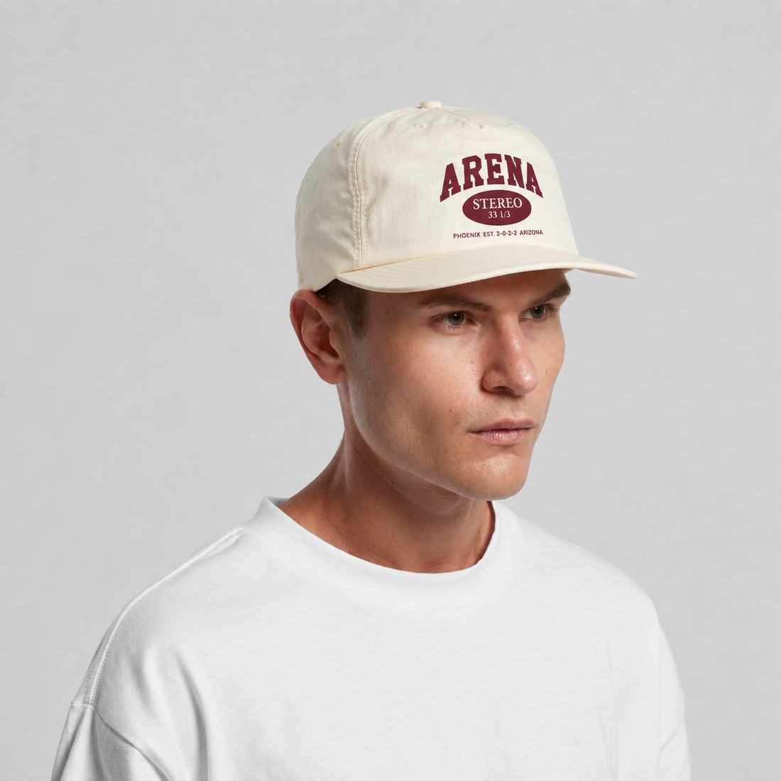 A man wearing an ecru-colored surf cap with a maroon 'ARENA STEREO' logo on the front, paired with a white crewneck t-shirt, against a light gray studio background.