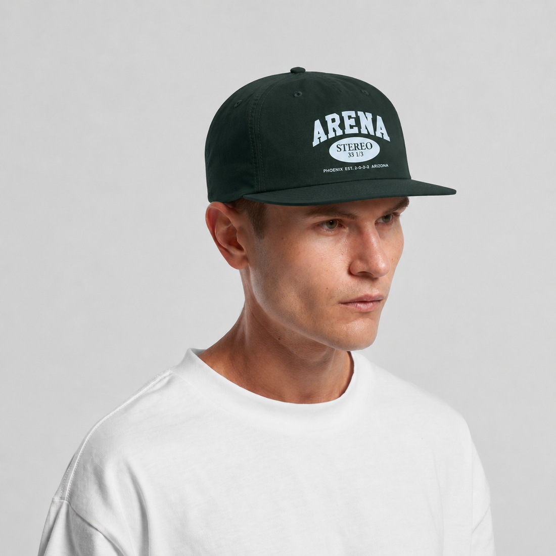 A man wearing a dark forest green snapback cap with a white 'ARENA STEREO' logo on the front, paired with a white t-shirt, against a light gray studio background.