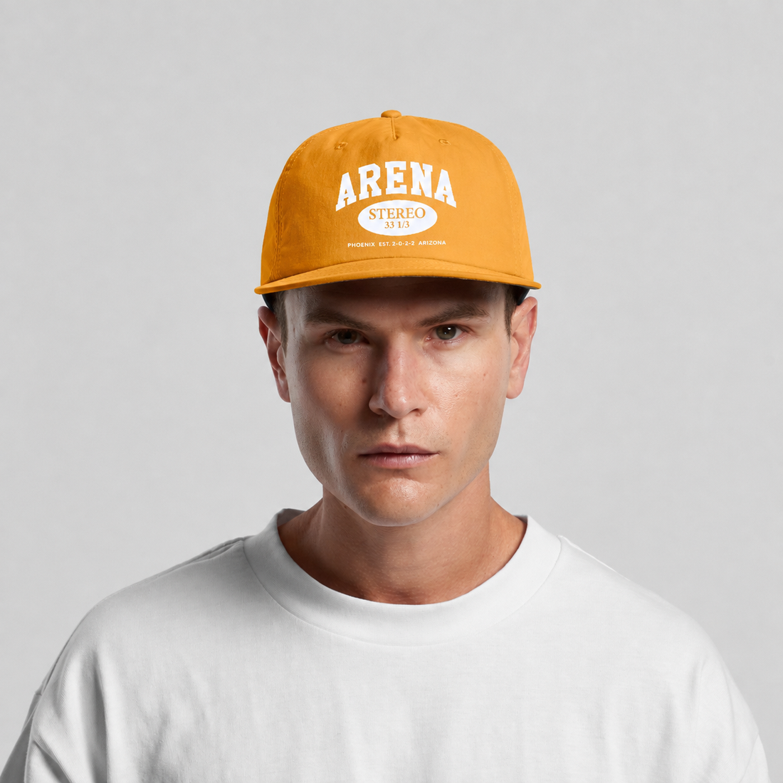 A man wearing a mustard yellow snapback cap with 'ARENA STEREO 2022' printed in white on the front, paired with a white t-shirt, against a light gray background.