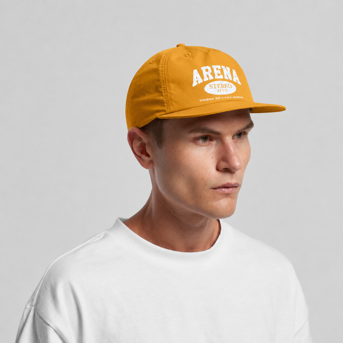 A man wearing a mustard yellow snapback cap with 'ARENA STEREO 2022' printed in white on the front, paired with a white t-shirt, against a light gray studio background.
