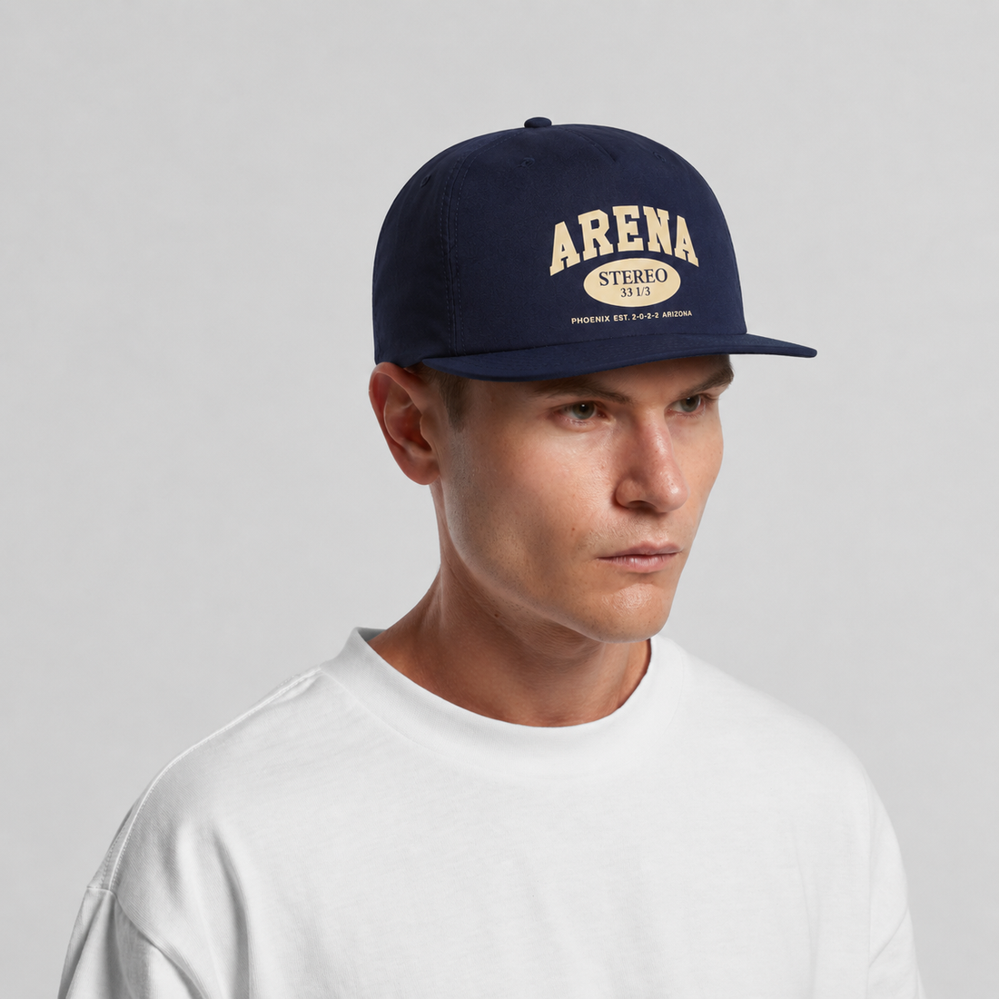 A man wearing a navy blue snapback cap with 'ARENA STEREO' printed in beige on the front, paired with a white t-shirt against a light gray background.