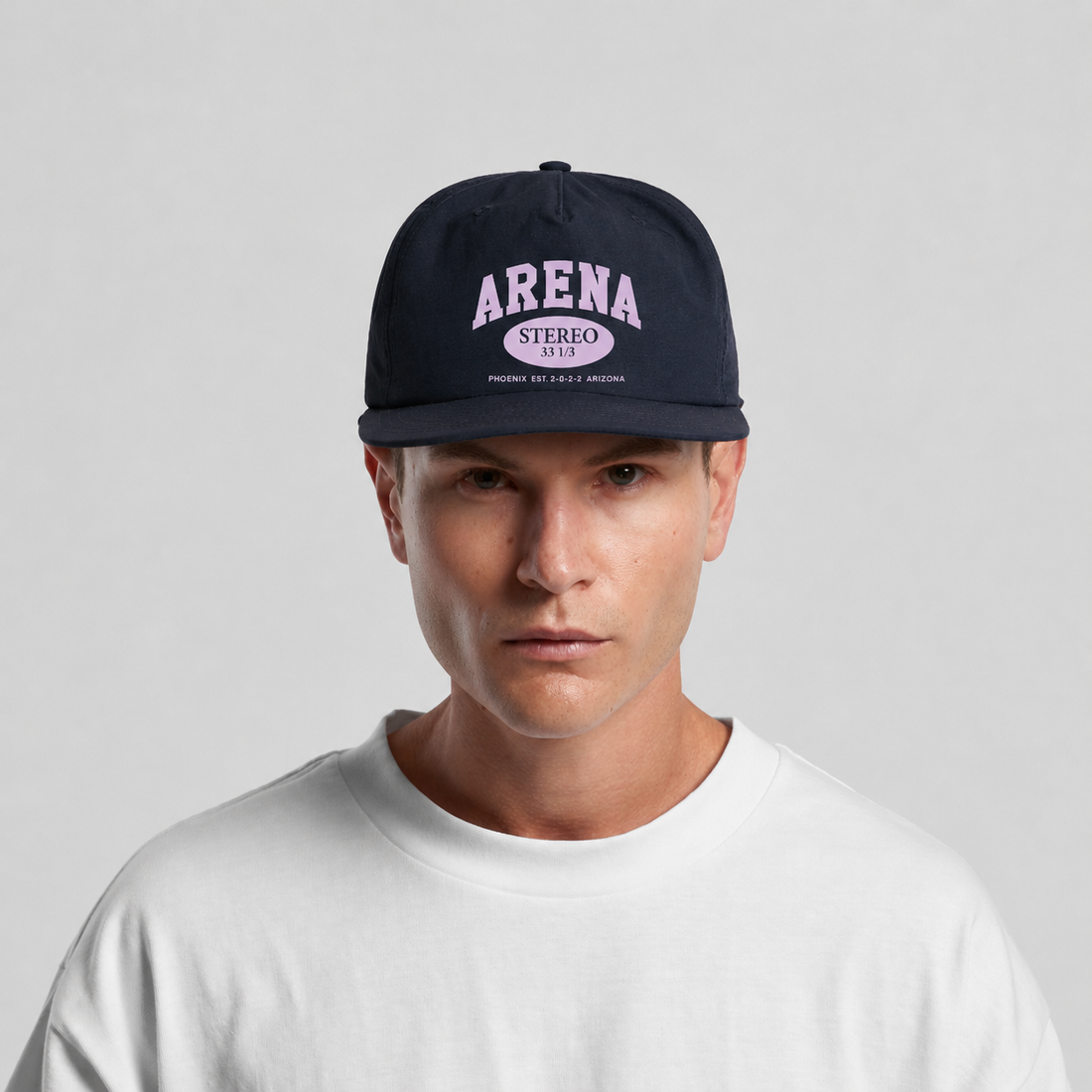 A man wearing a dark navy blue snapback cap with 'ARENA STEREO' printed in light purple on the front, paired with a white crewneck t-shirt, against a light gray studio background.