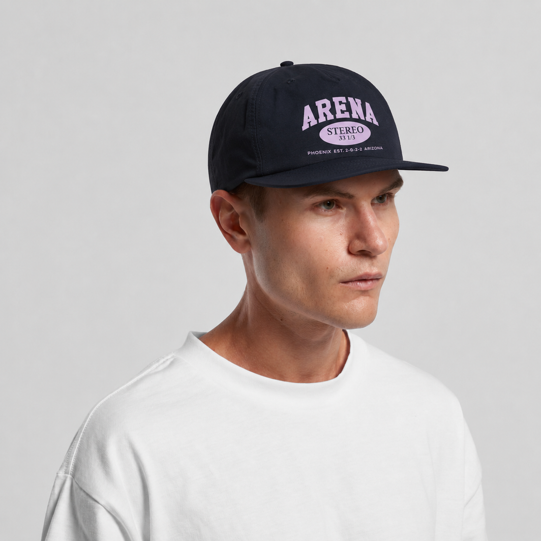 A man wearing a navy blue snapback cap with a purple and white 'Arena' logo, paired with a white crewneck t-shirt, photographed against a neutral light gray background.