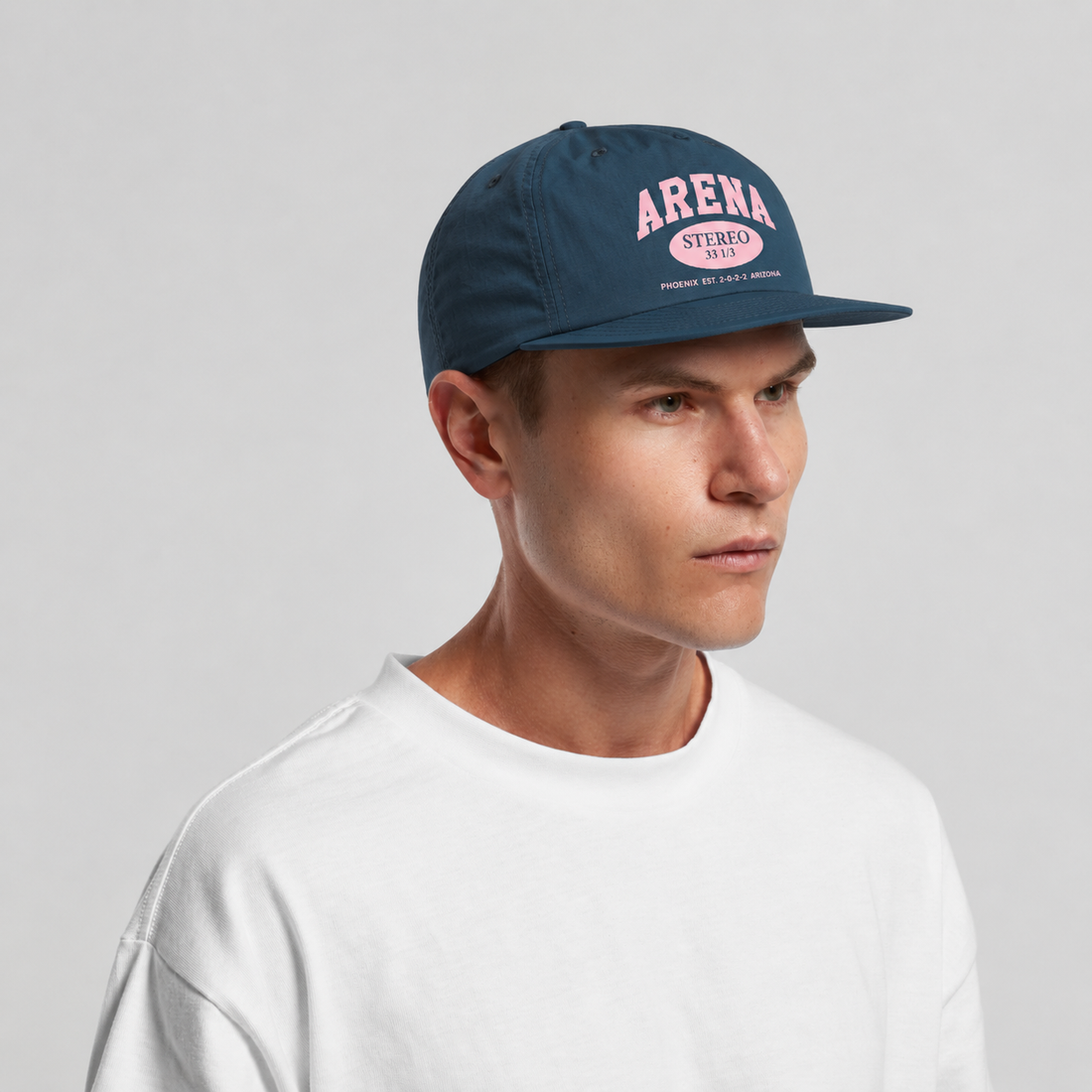 A man wearing a petrol blue Arena Stereo snapback cap and a white crewneck t-shirt, shown in a three-quarter profile against a light gray studio background.
