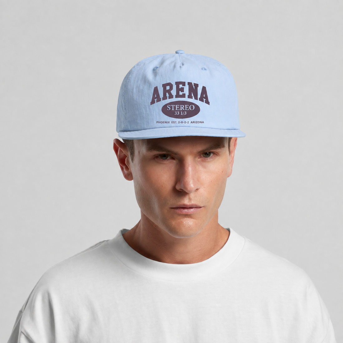 A man wearing a light blue snapback cap with 'ARENA STEREO 33 1/3' printed in dark red on the front, paired with a white t-shirt against a light gray background.