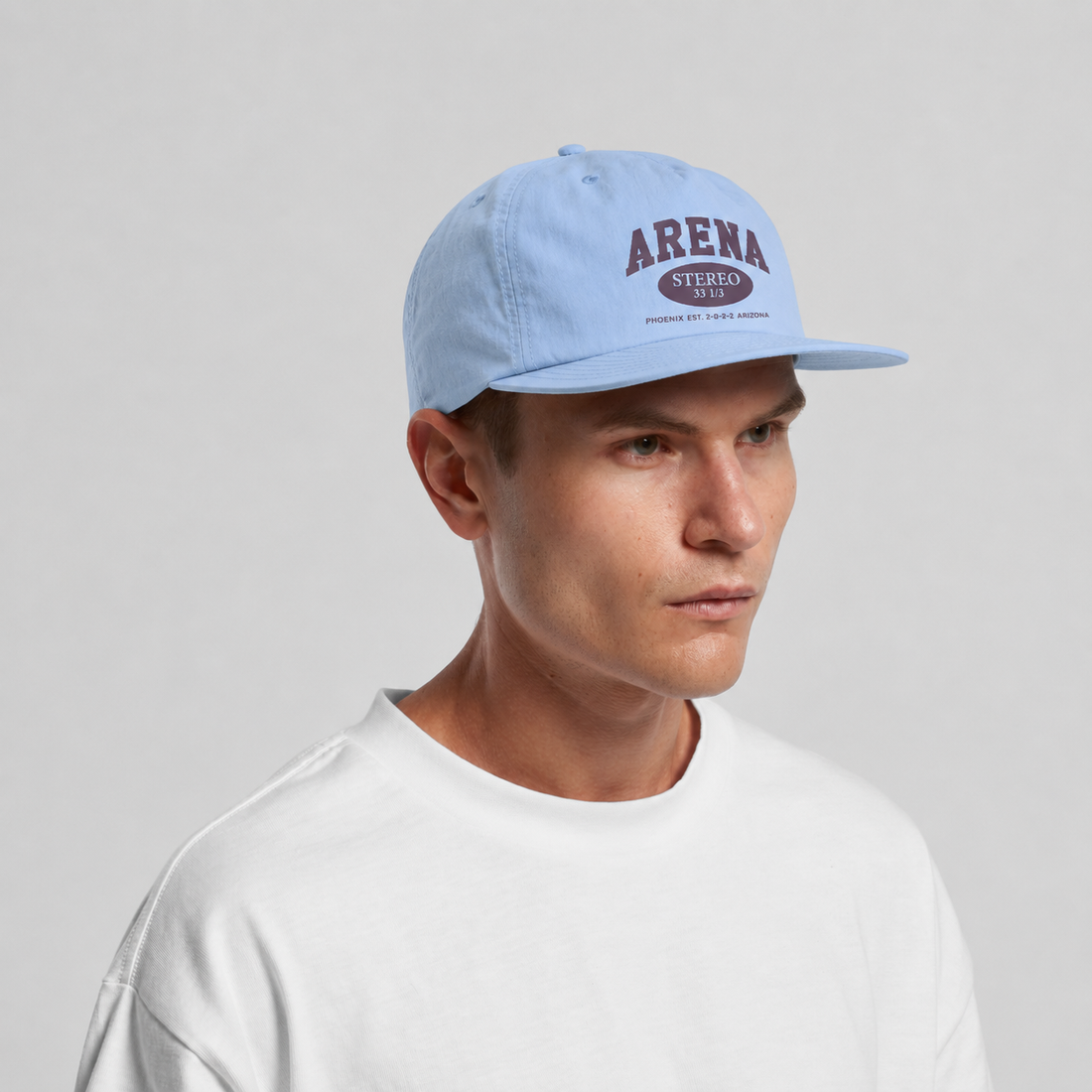 A man wearing a light blue flat-brim cap with a burgundy 'ARENA STEREO' logo on the front, paired with a white crewneck t-shirt, against a light gray studio background.
