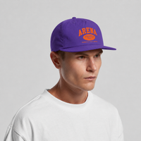 A man wearing a purple snapback cap with orange 'ARENA' and 'STEREO' text on the front, paired with a white t-shirt, against a light gray studio background.