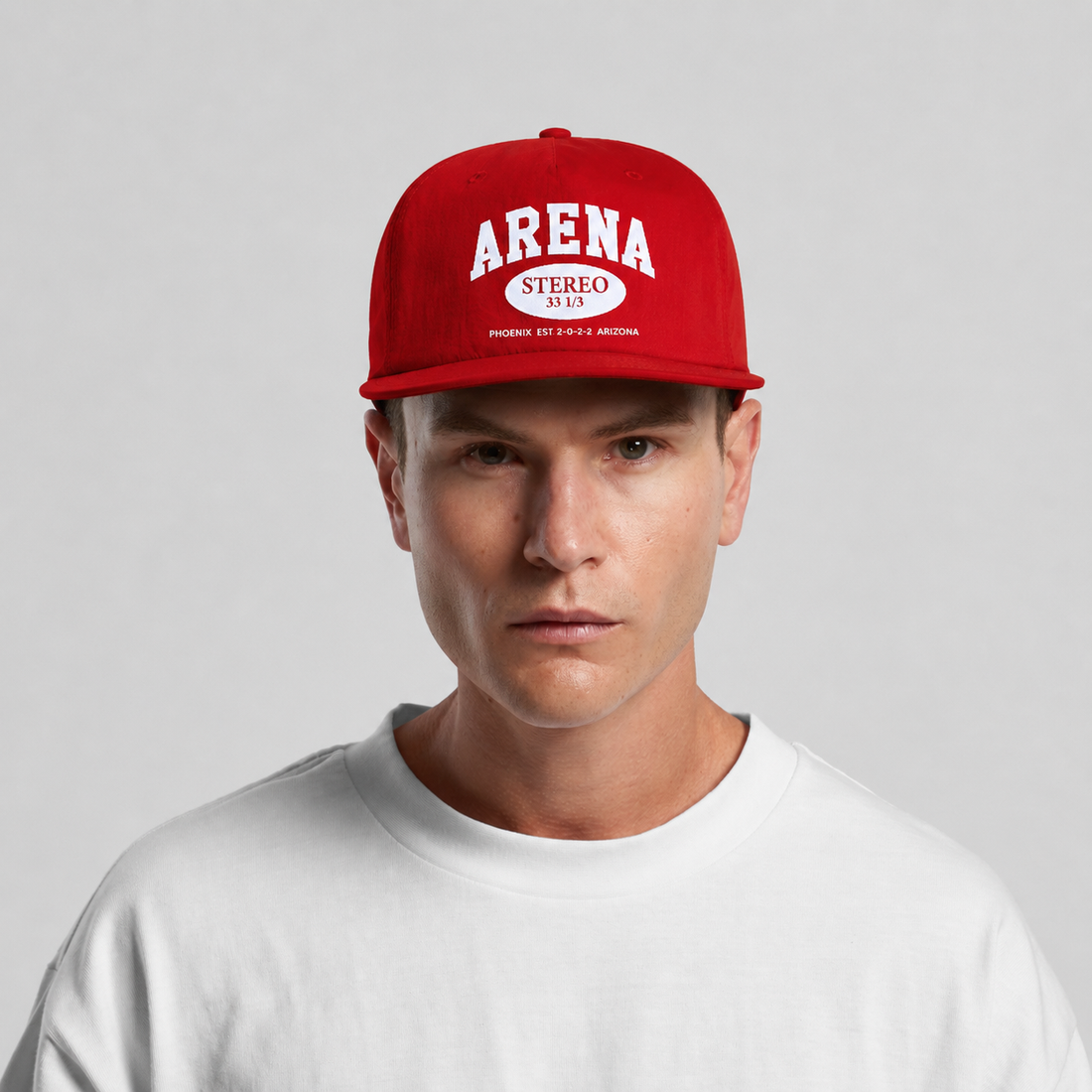 A man wearing a bright red snapback cap with 'ARENA STEREO' printed in white on the front, paired with a white crewneck t-shirt, against a light gray studio background.