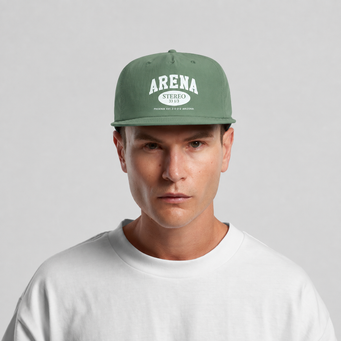 A man wearing a sage green snapback cap with a white 'ARENA STEREO' logo on the front, paired with a white crewneck t-shirt, against a light gray background.