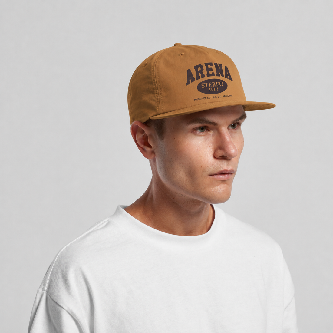 A man wearing a tan snapback cap with 'ARENA STEREO' printed in black on the front, paired with a white crewneck t-shirt, against a light gray background.